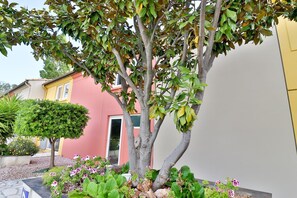 A building with a pink facade, a tree in the foreground, and a garden with various plants and flowers. - Hôtel Le Coudon (La Farlede)