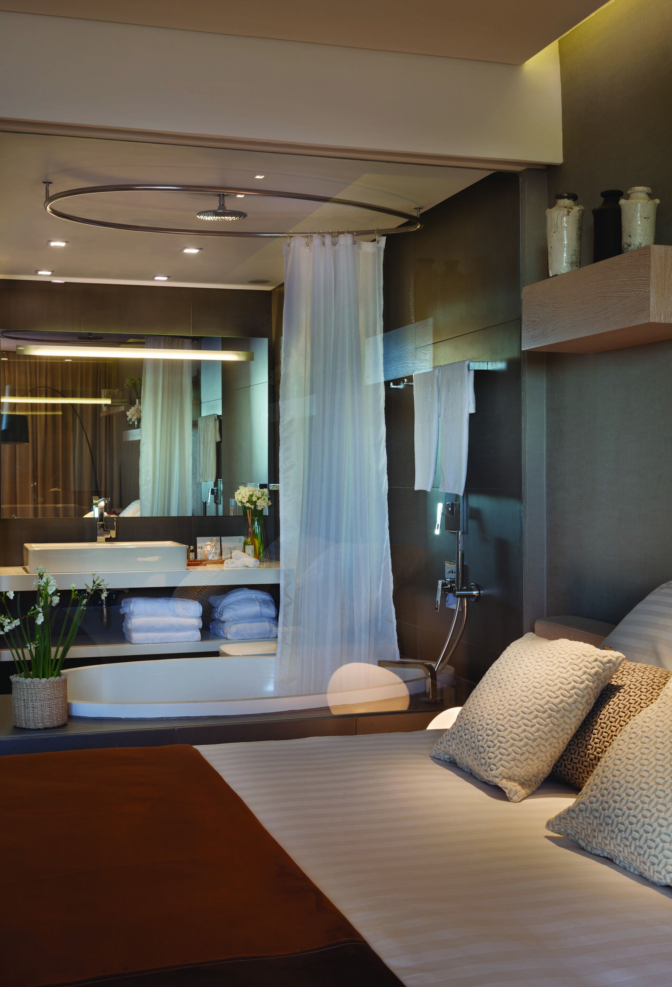 A modern bedroom with a large bed, a bedside table, a mirror, and a shower area.