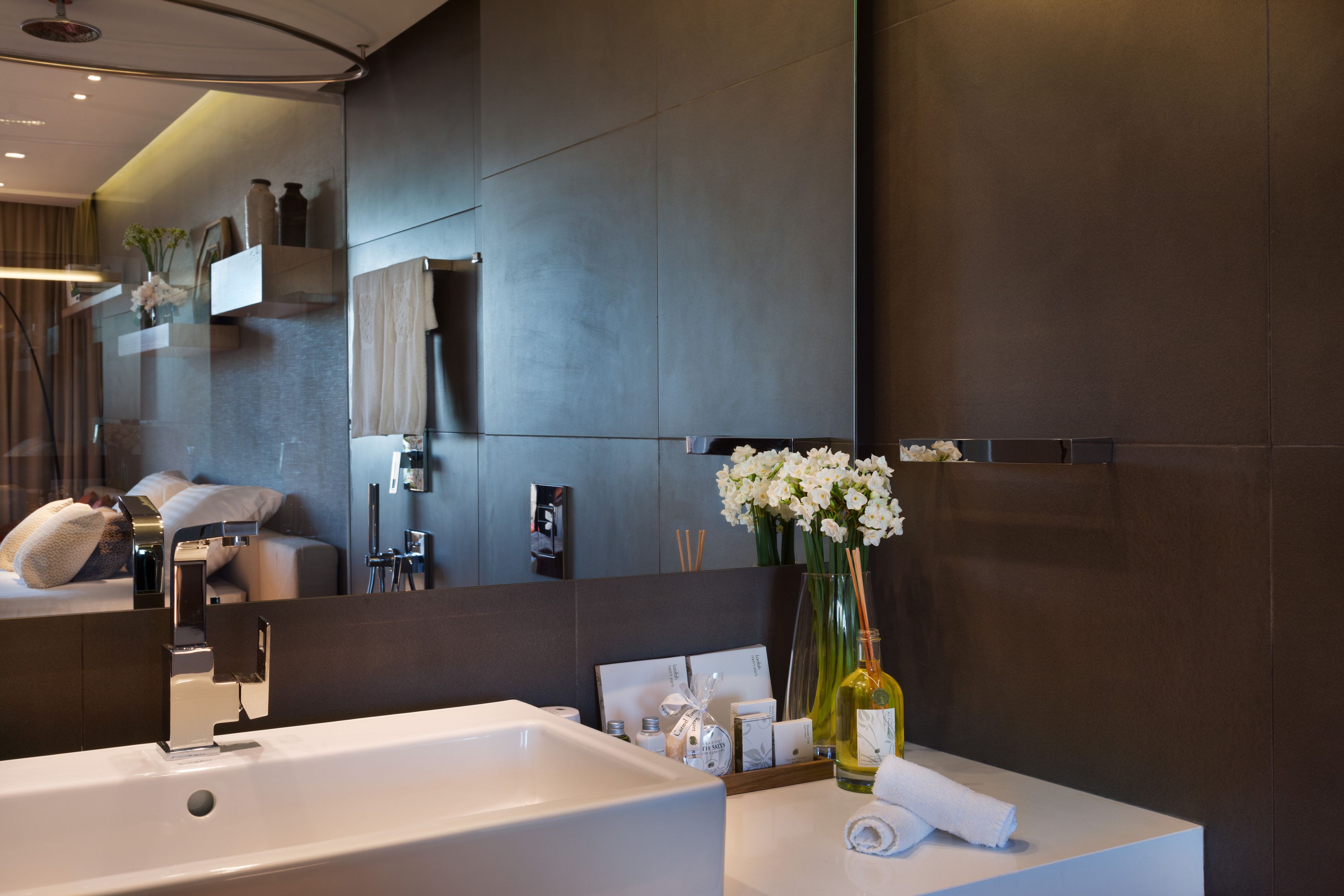 Un baño moderno con un espejo grande, un lavamanos blanco y un florero con flores blancas.