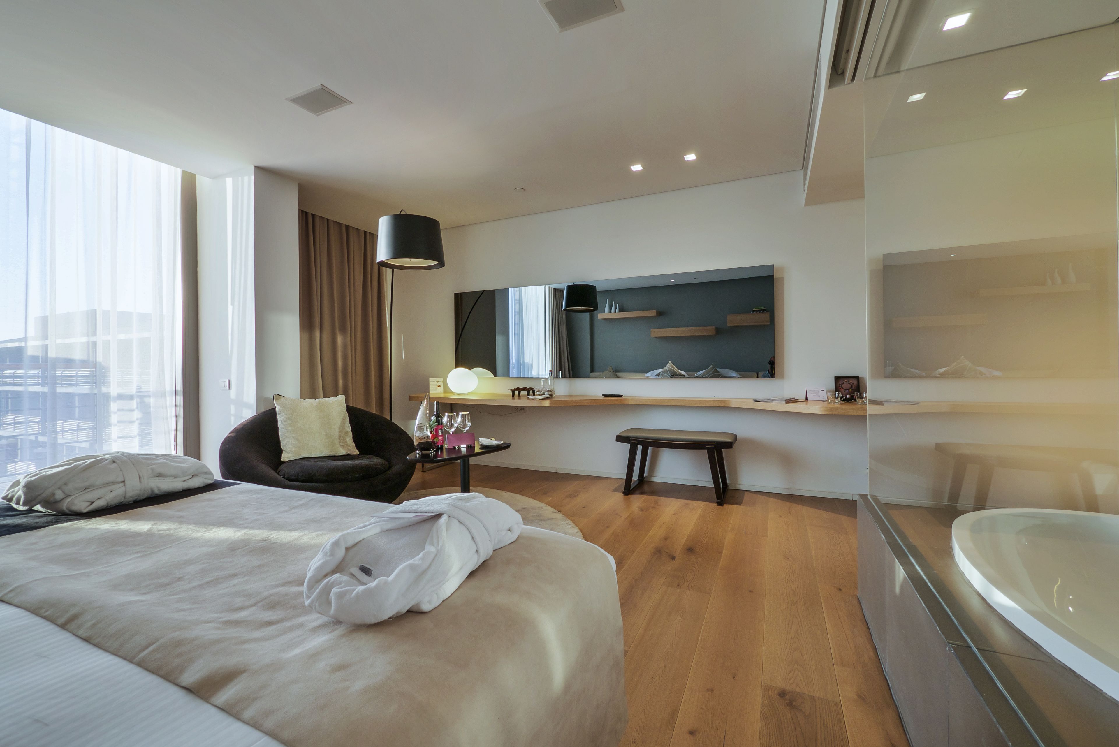 A modern hotel room with a large bed, a sitting area with a chair and table, a large mirror, and a glass-enclosed bathtub.