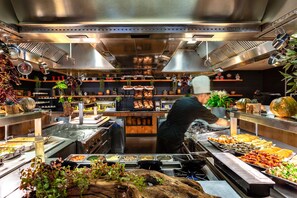 A commercial kitchen with stainless steel appliances, a chef preparing food, and various food items displayed on counters. - Cramim by Isrotel exclusive (Mateh Yehuda)