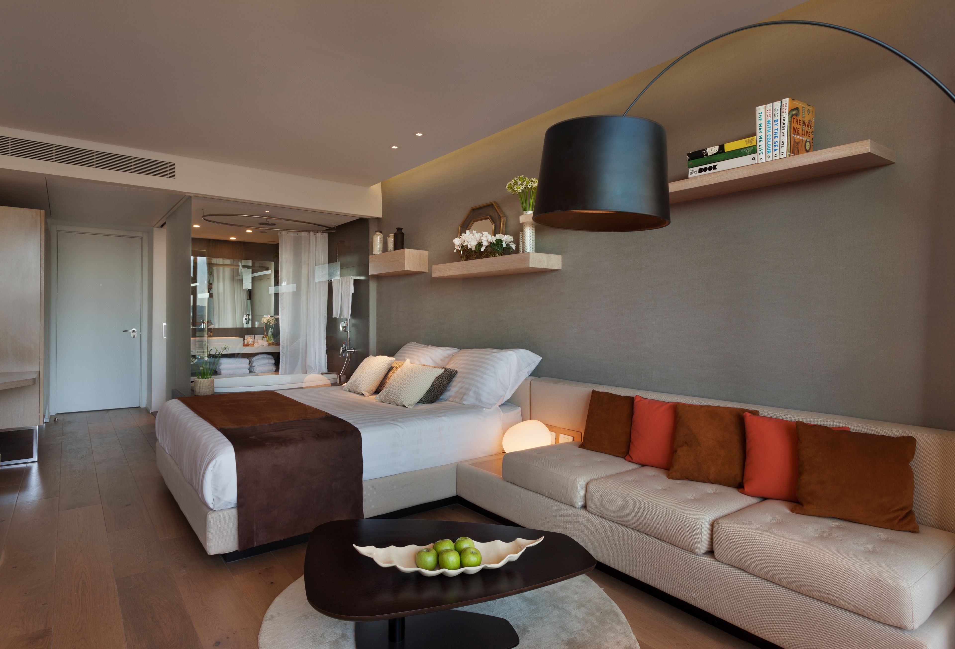 Ein modernes Hotelzimmer mit Bett, Sofa, einem Couchtisch mit Obst, einem Bücherregal und einer Pendelleuchte.