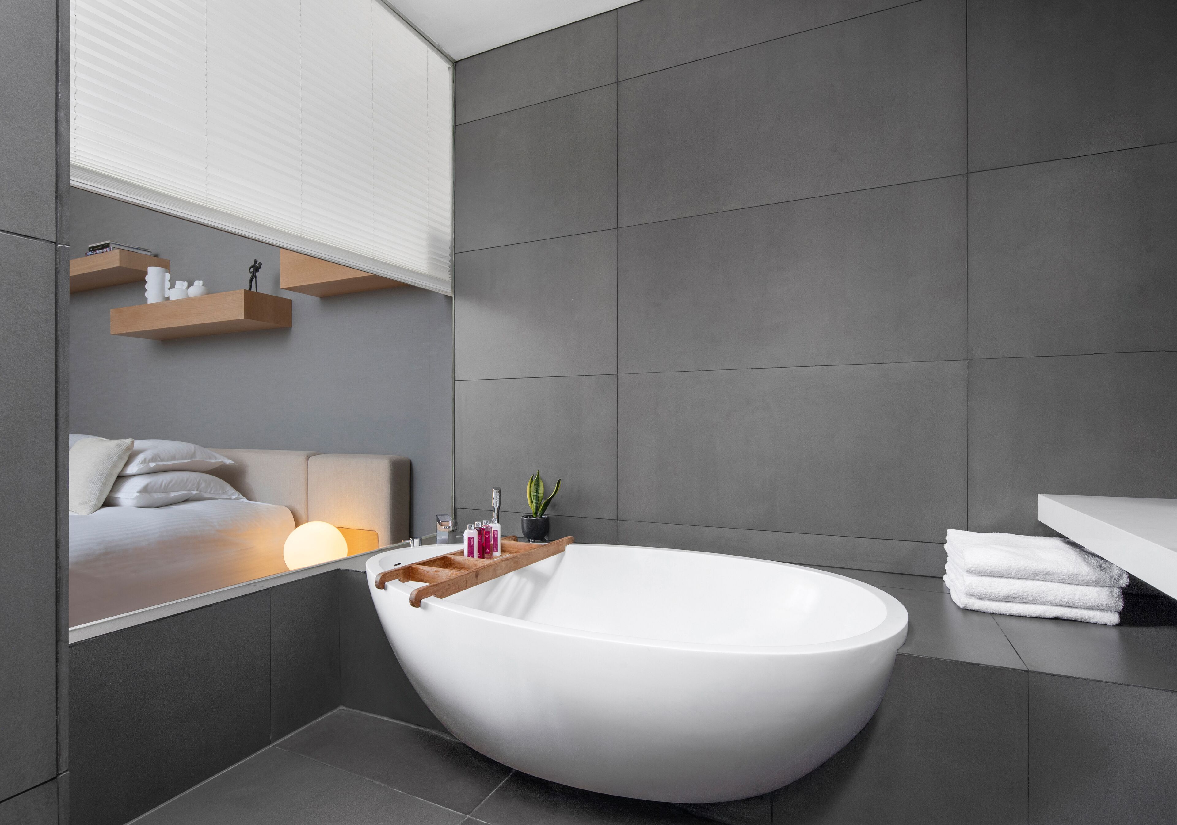 Un baño moderno con una bañera independiente, una mesita de noche con artículos de aseo y un espejo grande que refleja la habitación.