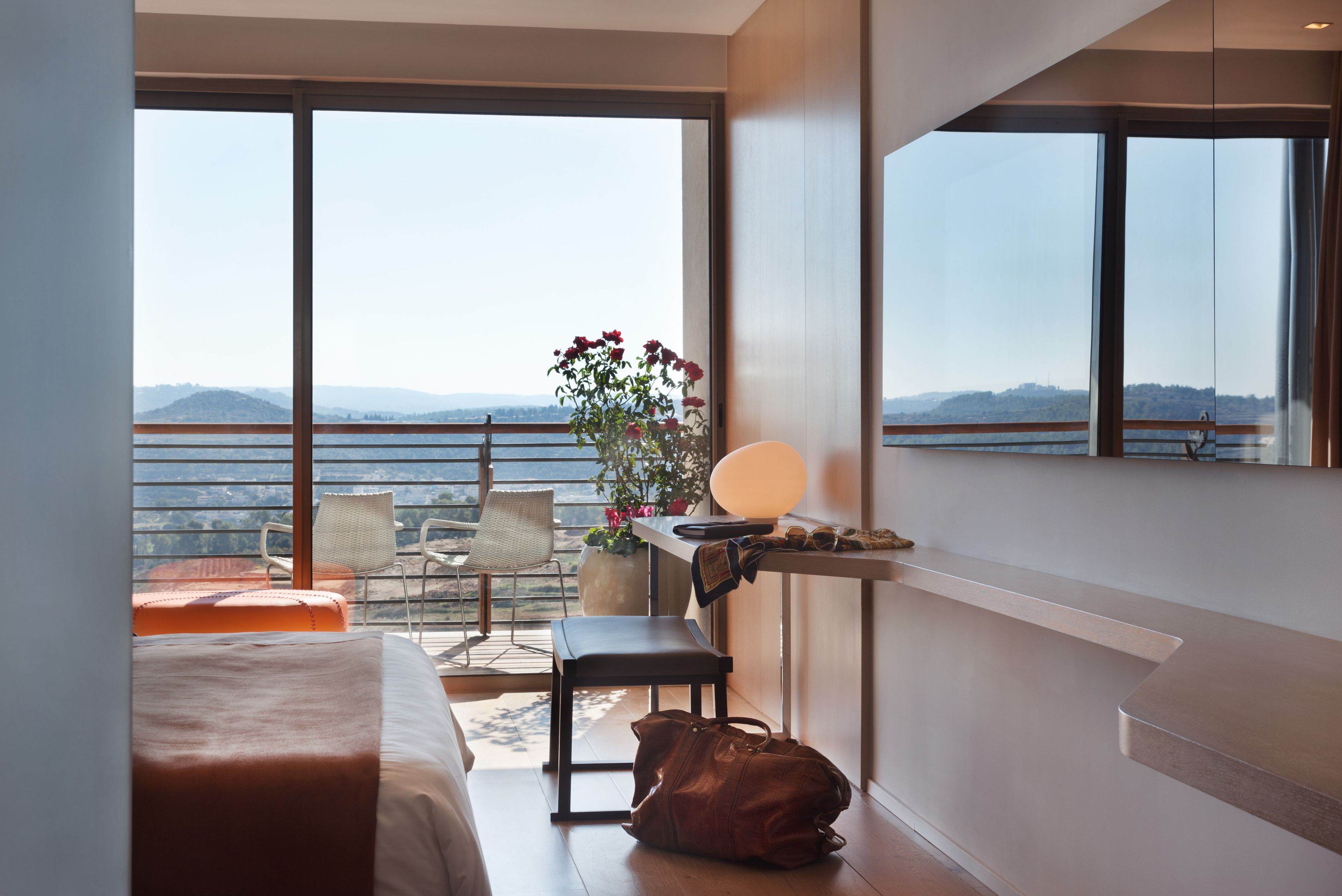 Habitación de hotel con cama, una silla, una mesita con lámpara, un banco y vistas a las montañas y al agua a través de amplias ventanas.