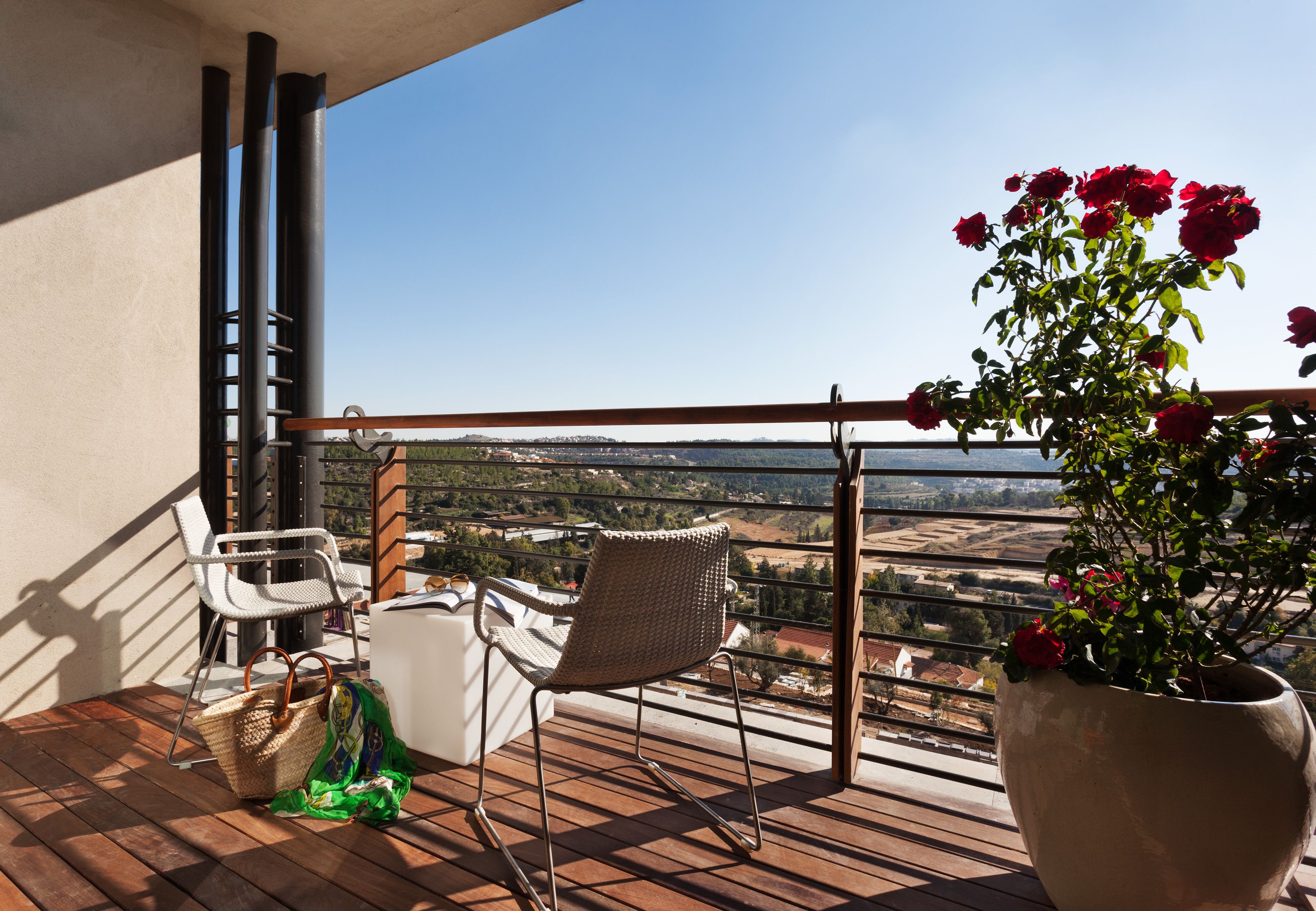 Un balcón con vistas a un valle, una silla de mimbre, una silla blanca, una maceta con flores rojas y una cesta de mimbre.