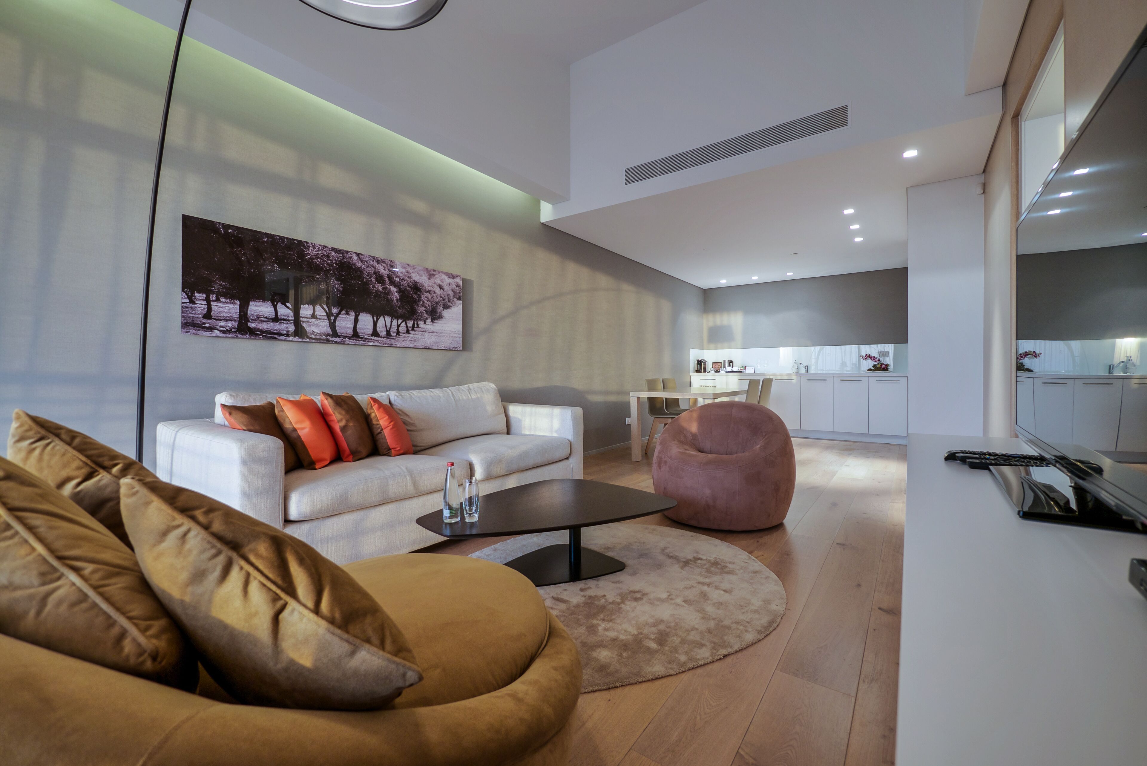 A modern living room with a sofa, a coffee table, and a pink chair. There is a kitchen in the background with white cabinets and a dining table.