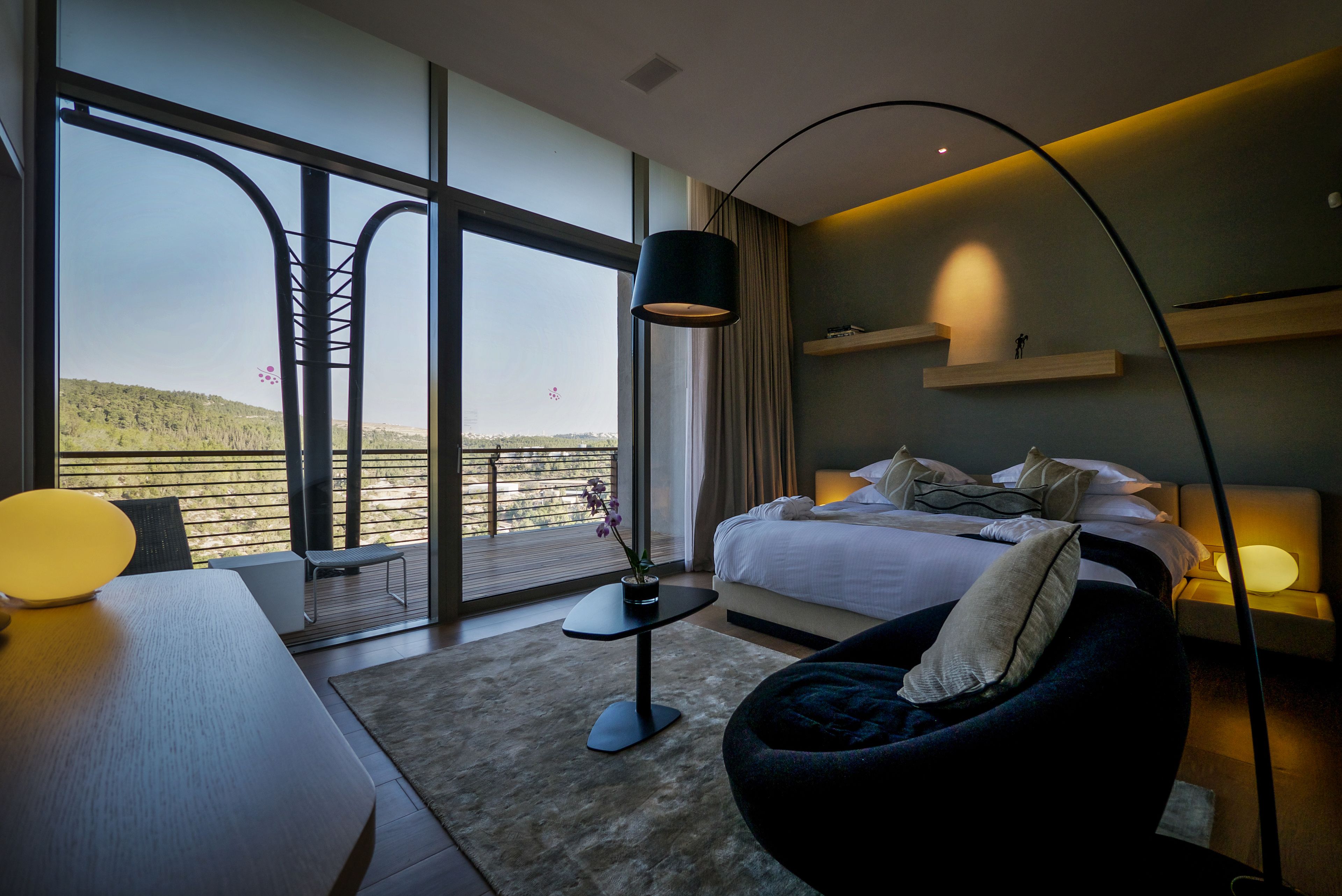 A modern hotel room with a large balcony, a bed with pillows, a desk with a lamp, and a chair.
