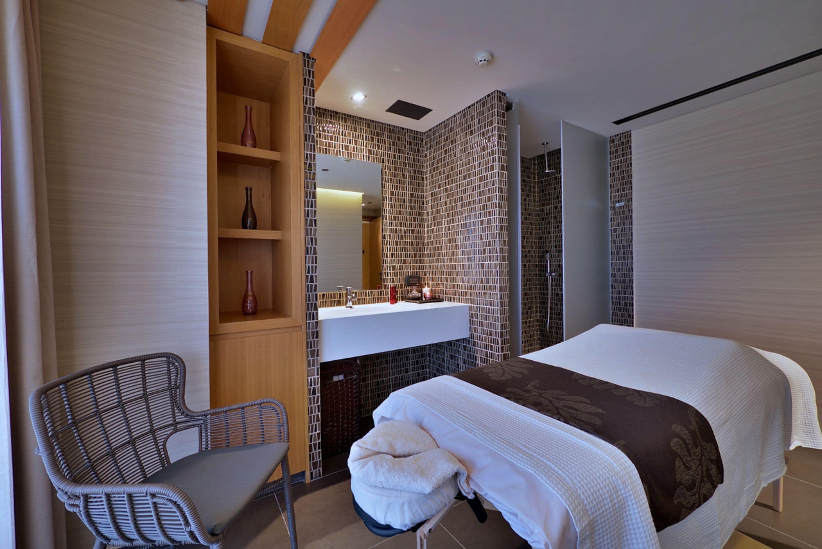 A modern hotel room with a large bed, a wooden chair, a vanity with a mirror, and a tiled wall.