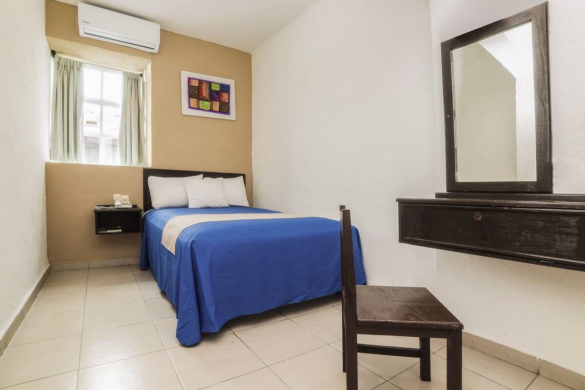A bedroom with a bed, a bedside table with a phone, a chair, a window with curtains, a wall-mounted mirror, and a colorful artwork.