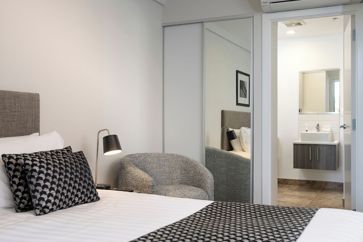 A modern hotel room with a bed, a chair, a desk, and a bathroom visible through a sliding door.