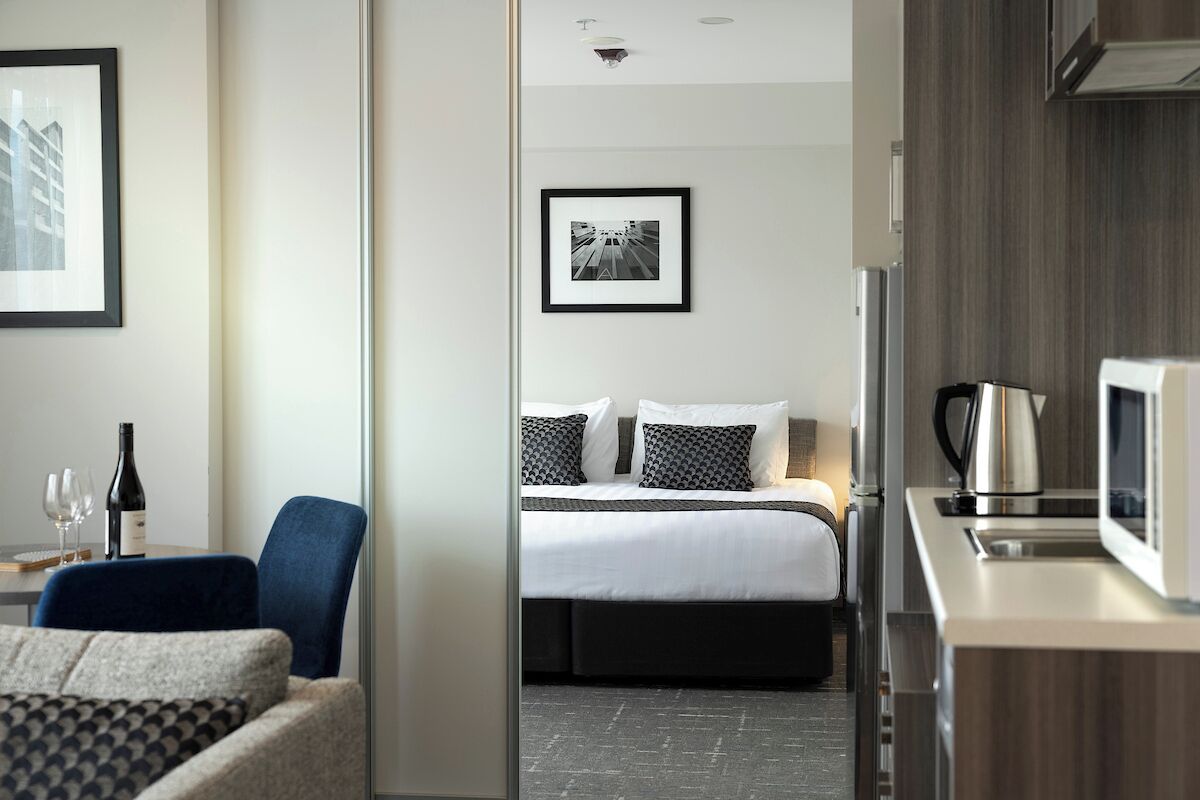 A modern hotel room with a bed, a seating area with a chair, a dining table with a bottle and glasses, a microwave, and a framed picture on the wall.