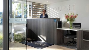 A reception desk with a 'QUEST' sign, a wheelchair-accessible sign, and a vase with flowers. - Quest on Beaumont (Auckland)