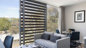 A modern living room with a grey sofa, two patterned cushions, a coffee table, and a view of a park and buildings through large windows. - Quest on Beaumont (Auckland)
