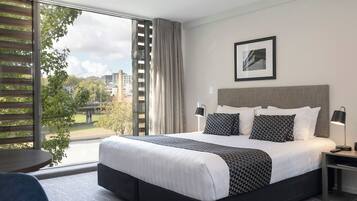 Habitación de hotel moderna con una cama grande, vista a través de una puerta corrediza de vidrio, un cuadro en la pared y una mesita con una lámpara.