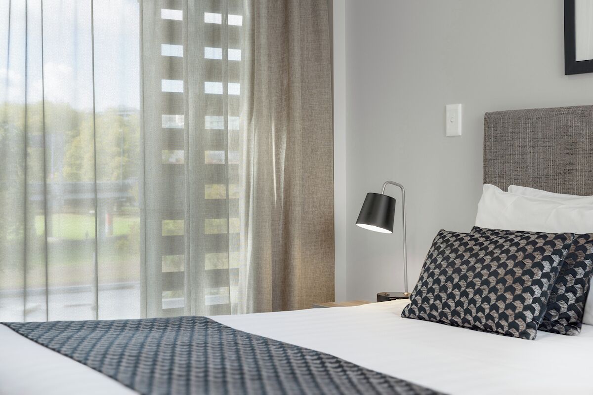 Une chambre d’hôtel avec un grand lit, une table de chevet, une lampe et une vue dégagée à travers des rideaux légers.