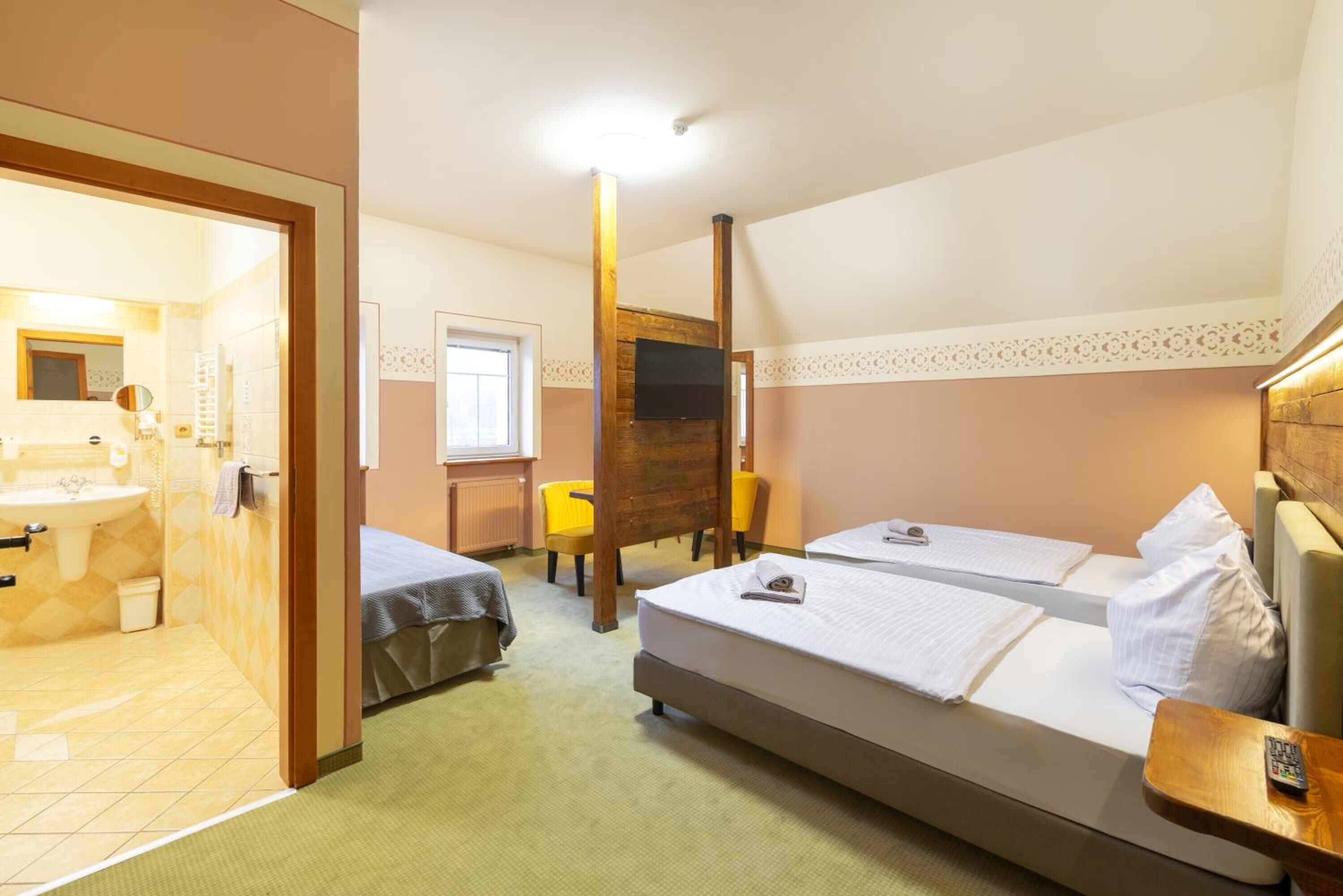 A hotel room with a bed, two bedside tables, a wooden chair, a small table, a TV, and a bathroom with a sink and mirror.