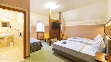 A hotel room with a bed, two bedside tables, a wooden chair, a small table, a TV, and a bathroom with a sink and mirror.
