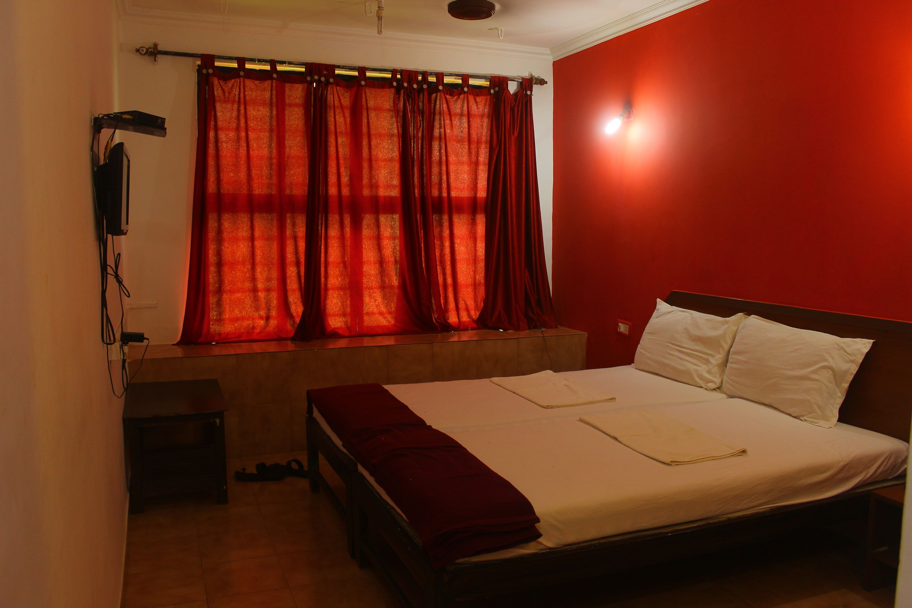 Quarto com cama, janela com cortinas vermelhas, mesa de cabeceira e aquecedor fixado na parede.
