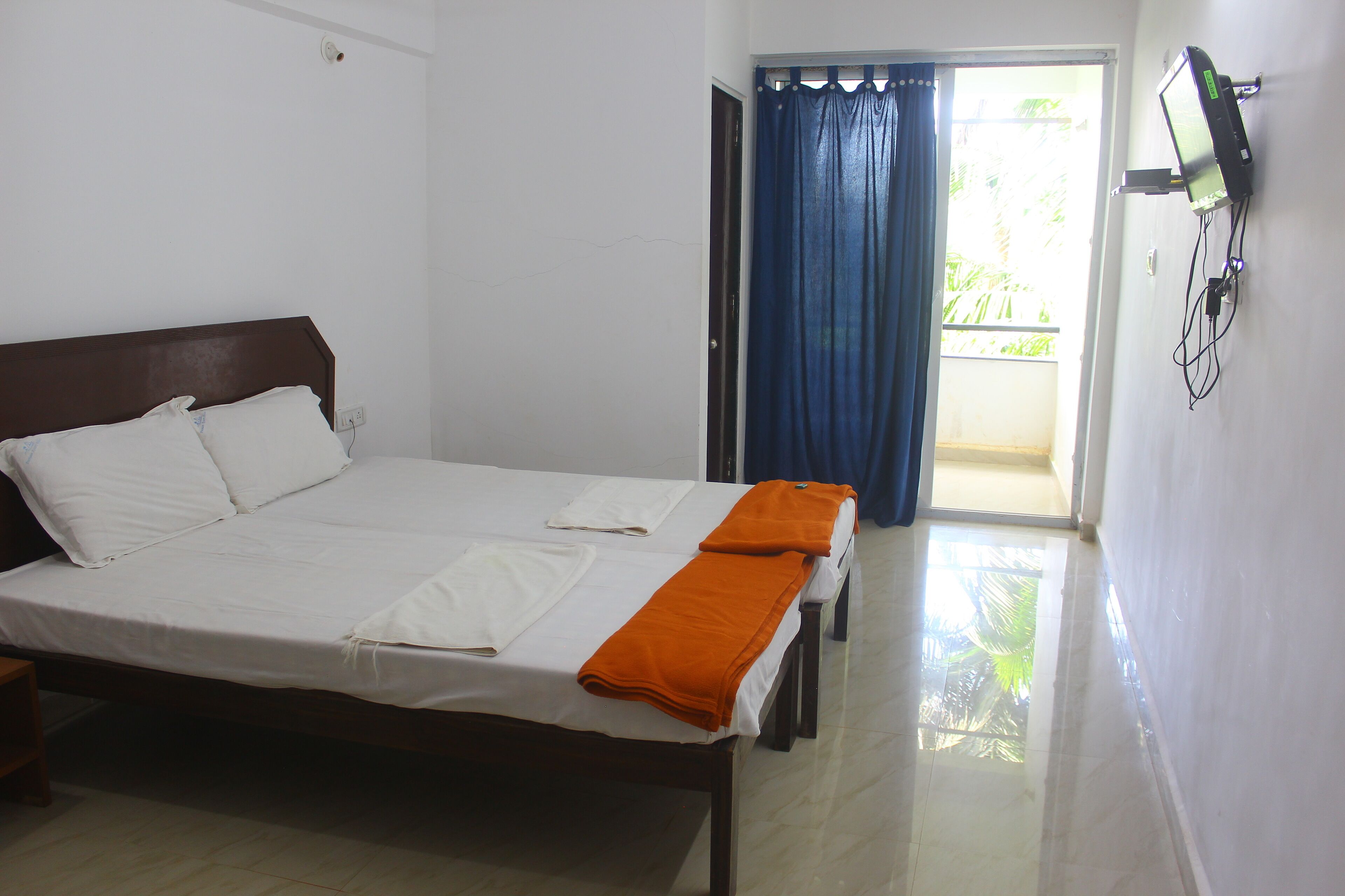 A neatly made bed with white linens and an orange towel, a television mounted on the wall, and a door leading to a balcony with blue curtains.