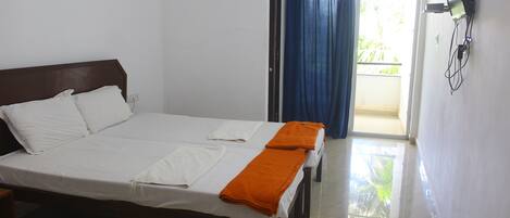 A neatly made bed with white linens and an orange towel, a television mounted on the wall, and a door leading to a balcony with blue curtains.