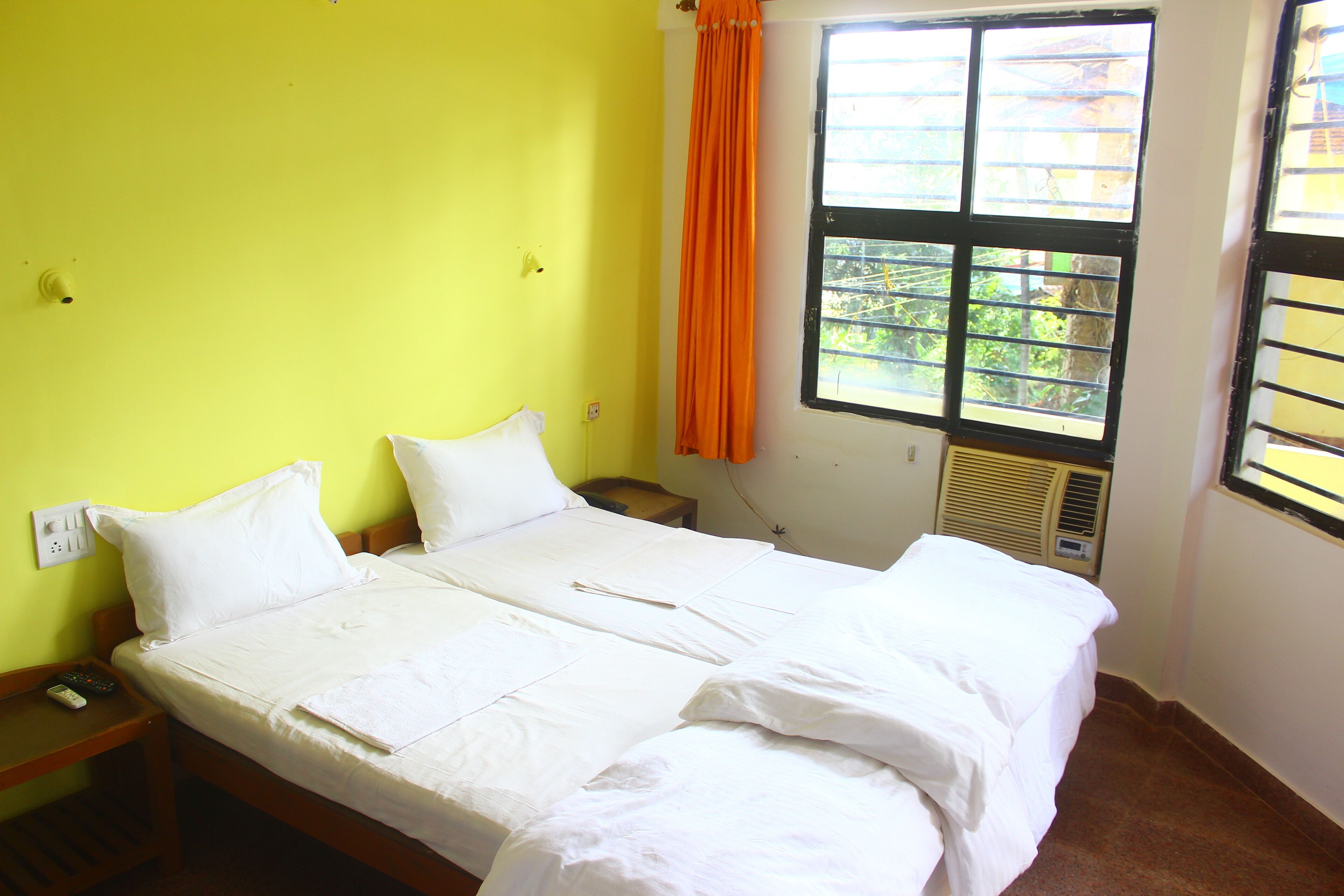 Ein Doppelbett mit weißer Bettwäsche, ein Nachttisch aus Holz, eine Fernbedienung und ein Fenster mit orangefarbenem Vorhang.