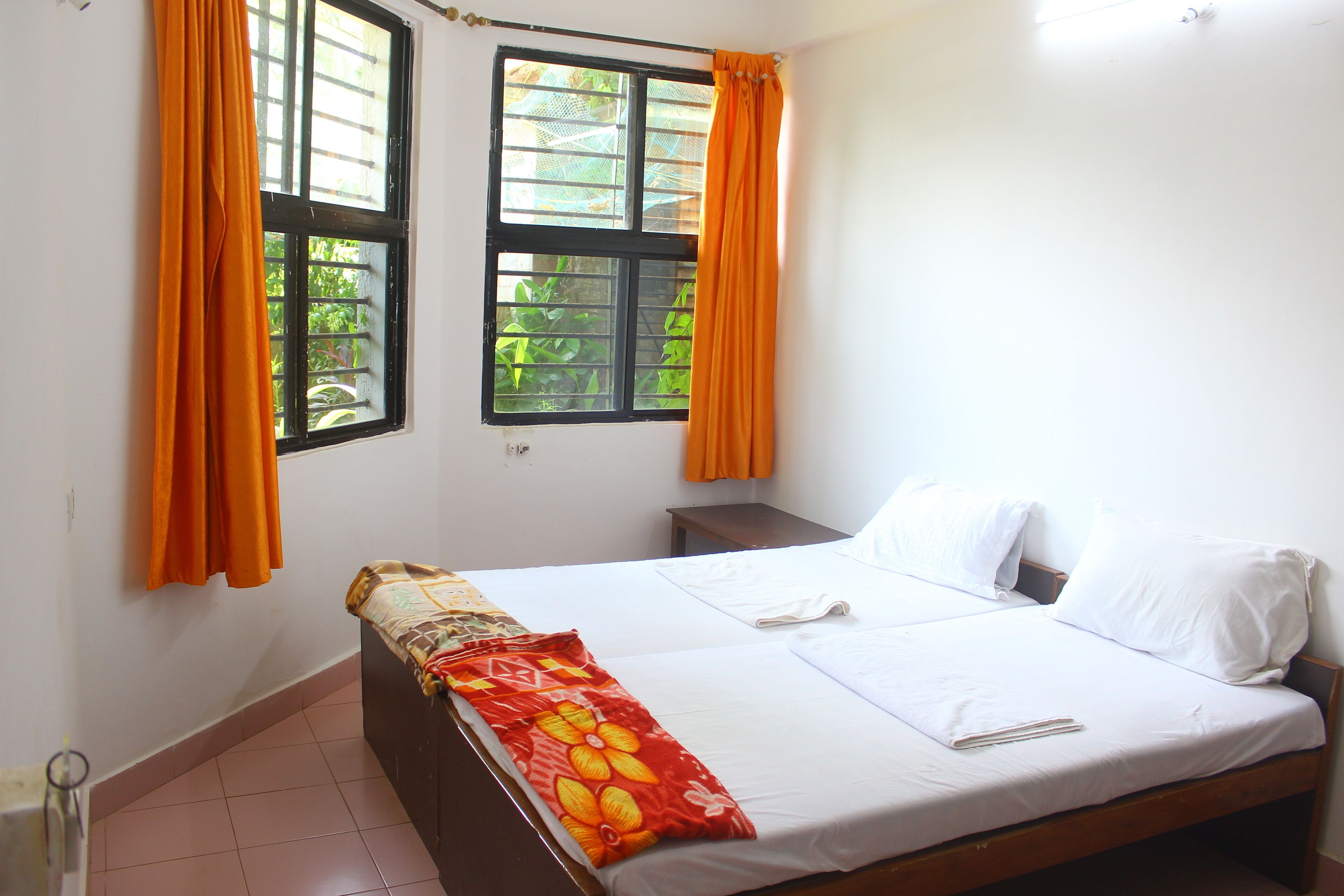 A bedroom with two beds, orange curtains, and a window with a view of greenery.