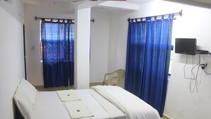 Una cama individual con ropa blanca, una silla, un televisor montado en la pared y cortinas azules.