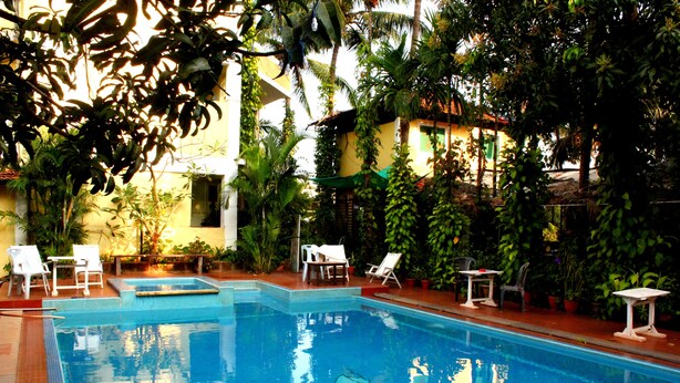 Una piscina con sillas de descanso y mesas, rodeada de vegetación y un edificio al fondo.