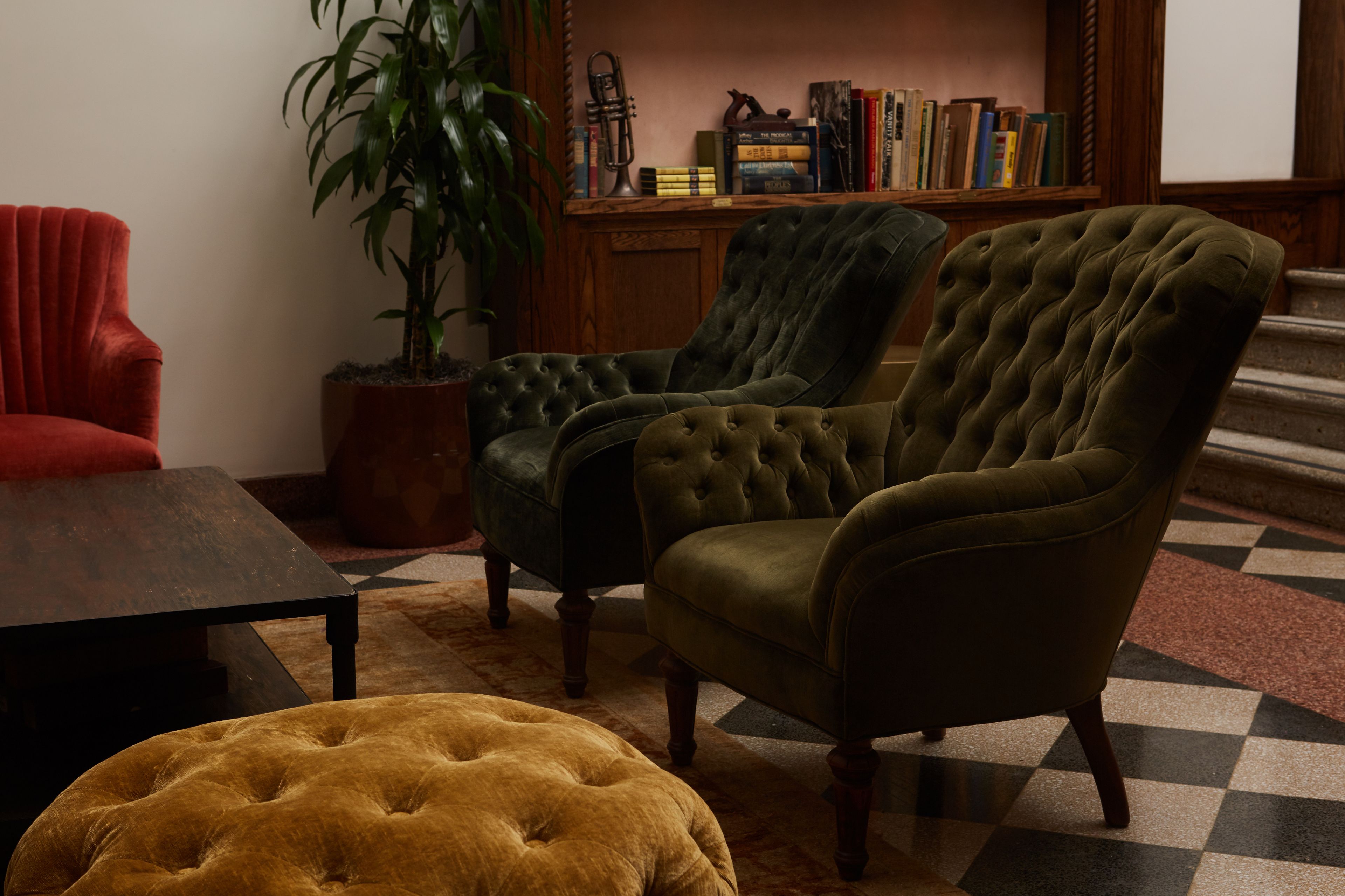 A room with a checkered floor, a red armchair, a green armchair, a brown ottoman, and a bookshelf with books and a plant.