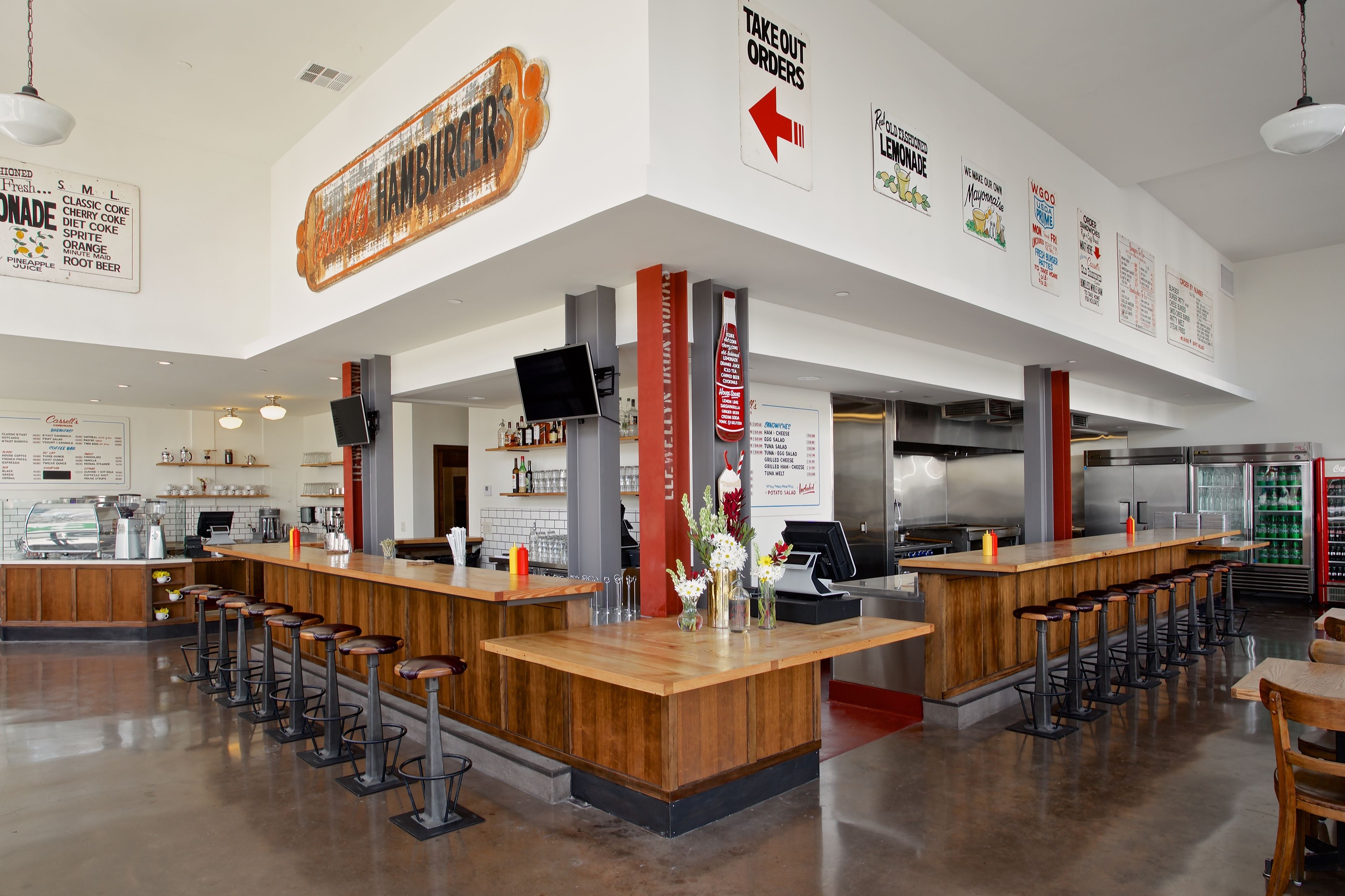 A modern bar area with wooden counters and stools, a large menu board, and various beverage dispensers.