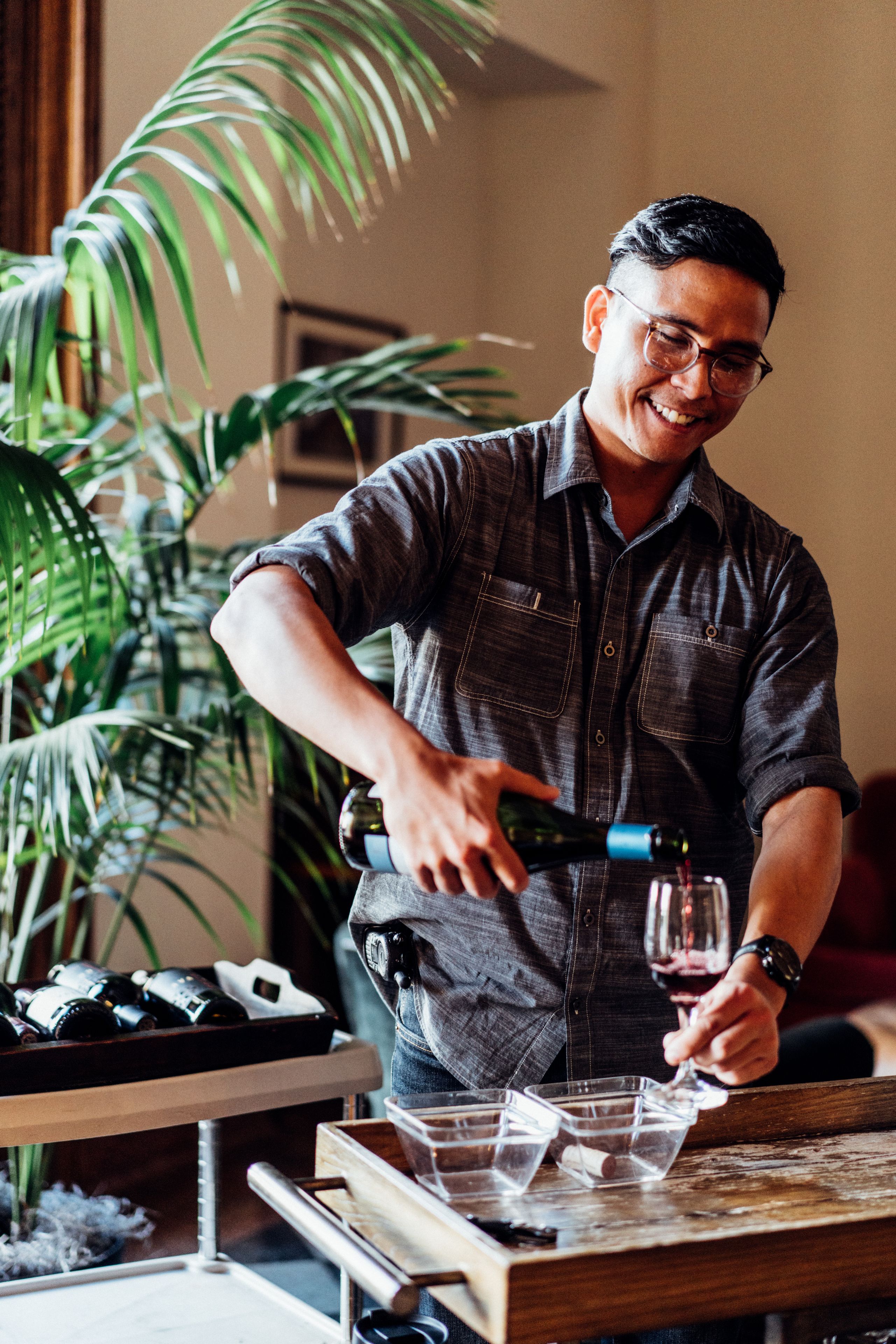 A person pouring wine into a glass on a wooden table with a plant in the background.