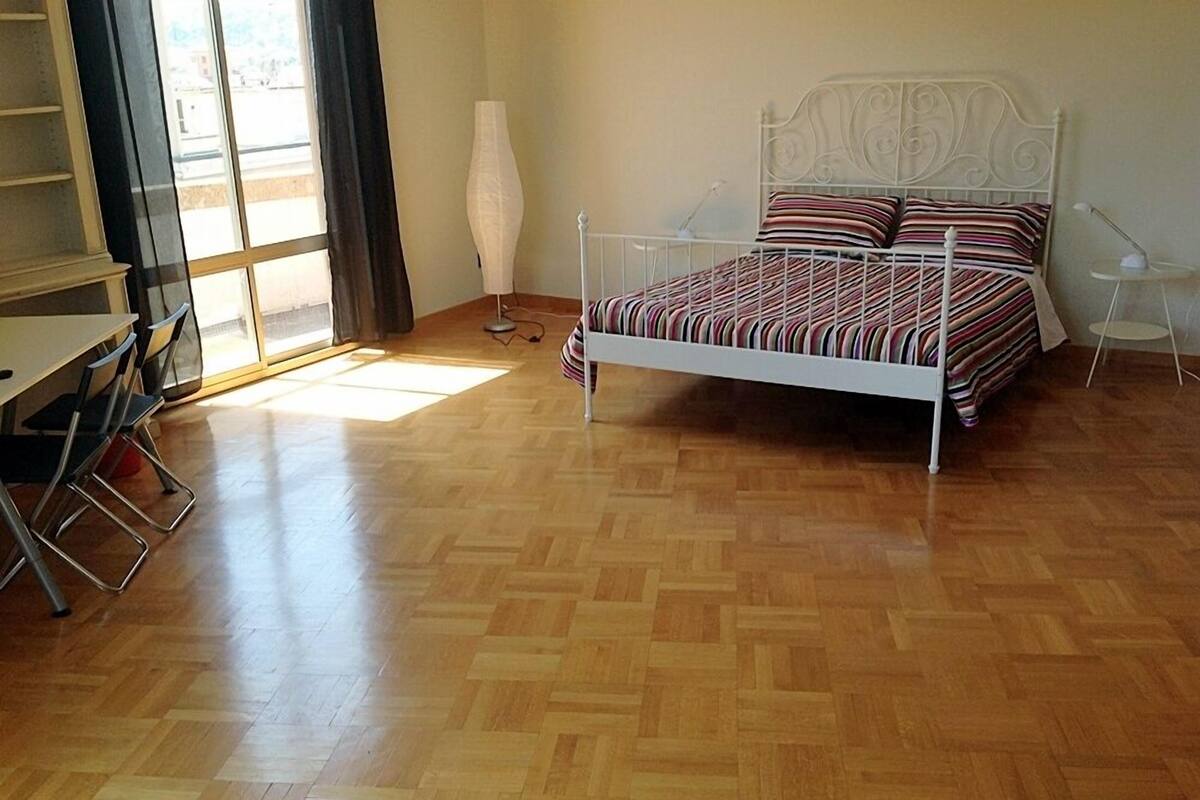 A bedroom with a wooden parquet floor, a white metal bed frame, striped bedding, a standing lamp, and a window with curtains.