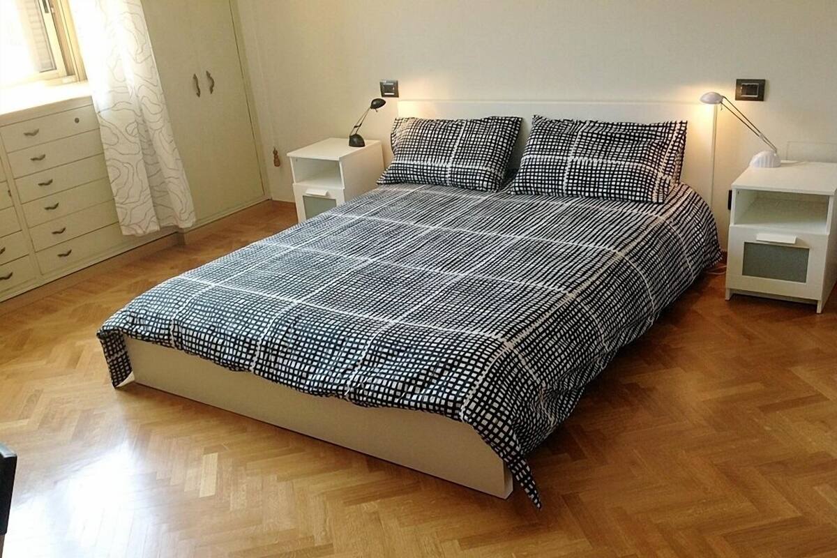 A bedroom with a bed, two bedside tables, a nightstand, a dresser, and a window with curtains.