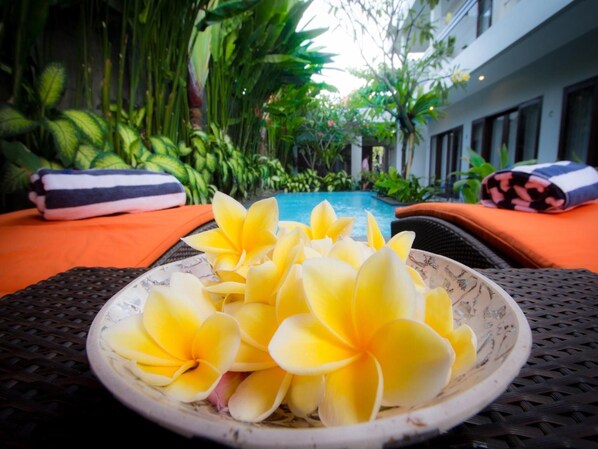 A bowl of yellow flowers on a table with a pool and greenery in the background.