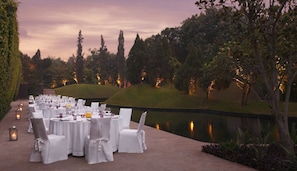Un área de comedor al aire libre con mesas redondas, manteles blancos y sillas, frente a un fondo de árboles y una masa de agua, iluminada con luces ambientales.