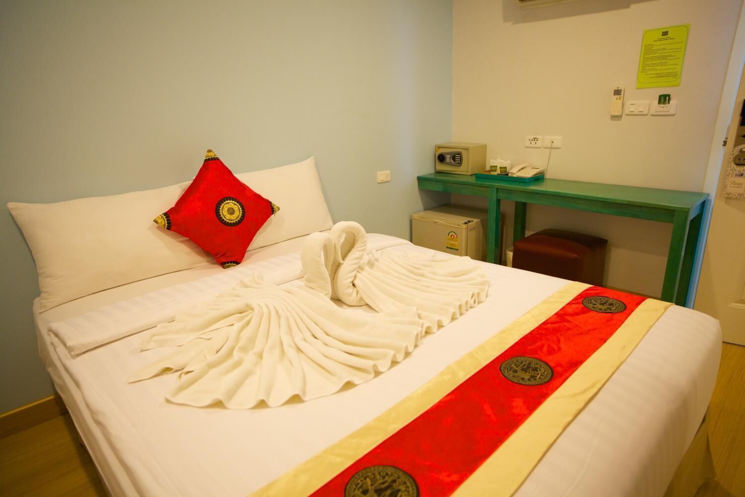A hotel room with a neatly made bed, a red pillow, a green desk with a telephone and a box, and a wall-mounted light switch.