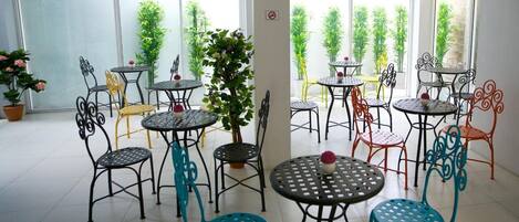 A spacious indoor area with multiple round tables and chairs, surrounded by large windows with greenery outside.