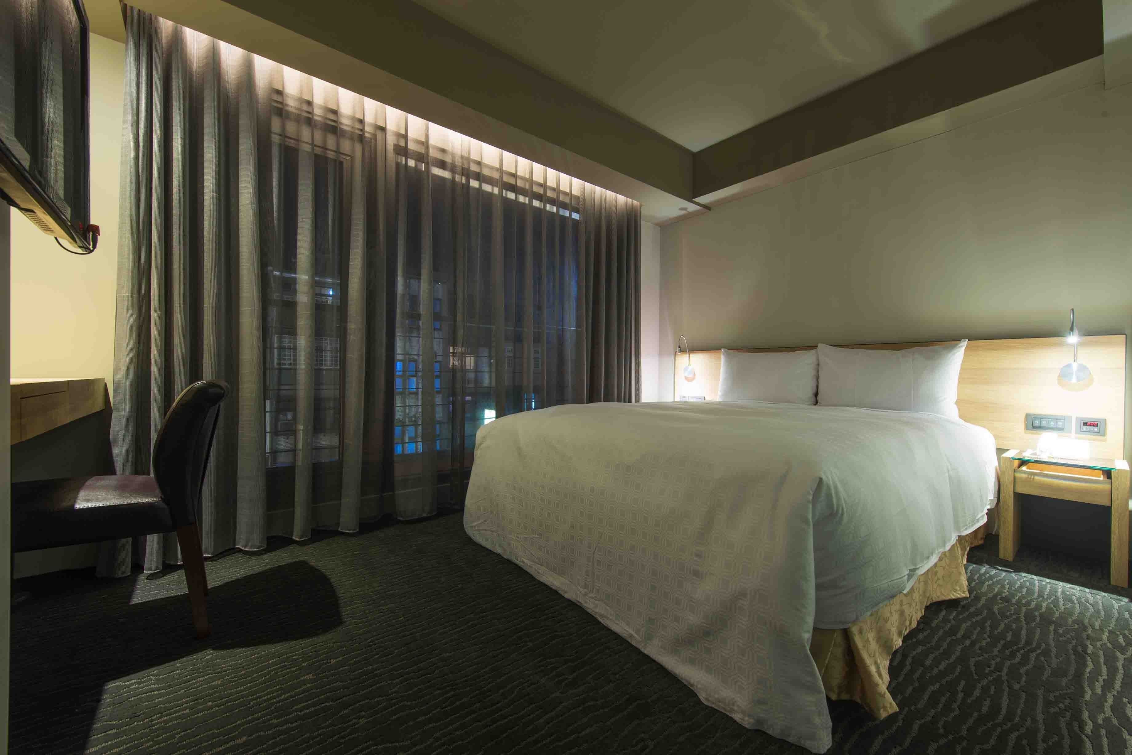 A hotel room with a large bed, a desk, a chair, and a window with curtains.