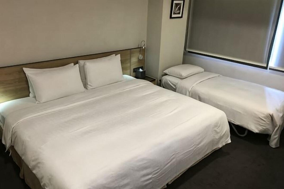 A hotel room with a large bed, a smaller bed, a bedside table, a lamp, and a window.