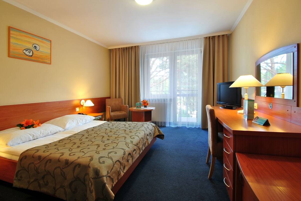 Une chambre d’hôtel comprenant un lit, un bureau avec un ordinateur, une chaise, une petite table avec une lampe et une fenêtre avec des rideaux.