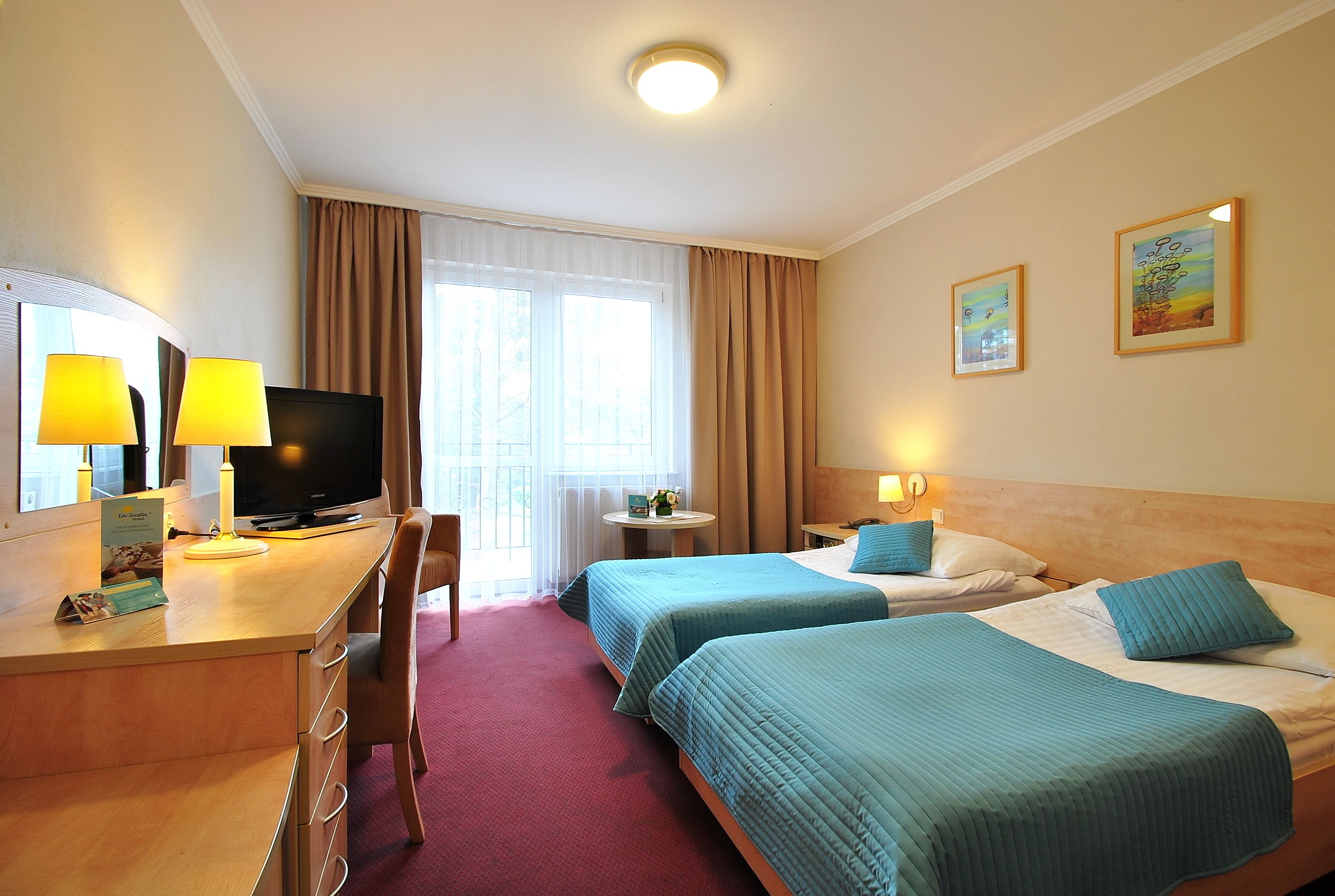 A hotel room with two beds, a desk with a computer and lamp, a TV, and a window with curtains.