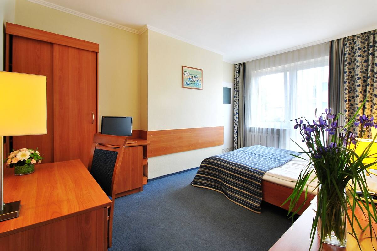 Une chambre d’hôtel comprenant un lit, un bureau avec un ordinateur, une chaise, une télévision, une fenêtre avec des rideaux et un vase avec des fleurs.