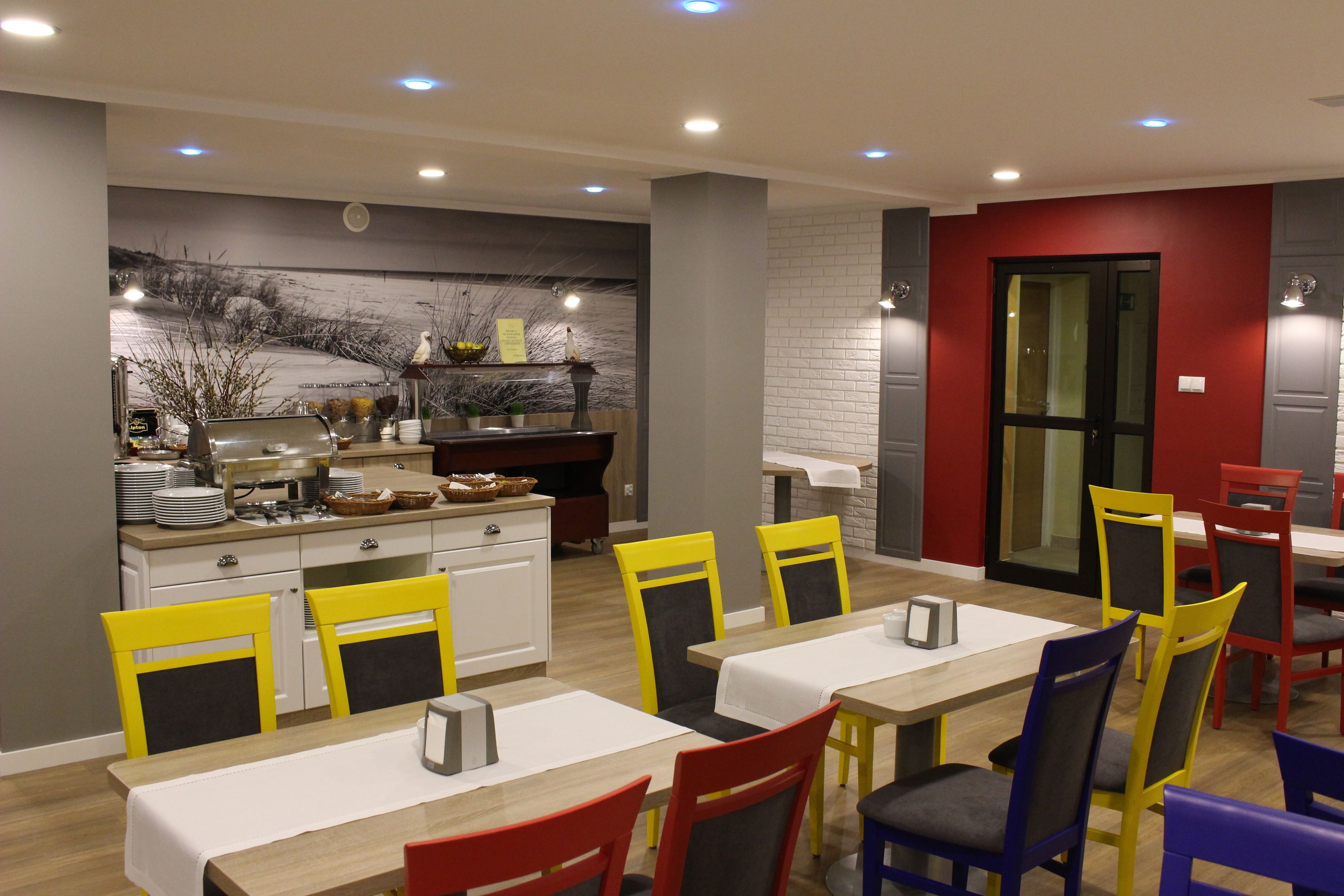 A modern dining area with a long table and multiple chairs, a kitchen area with a buffet, and a red accent wall.