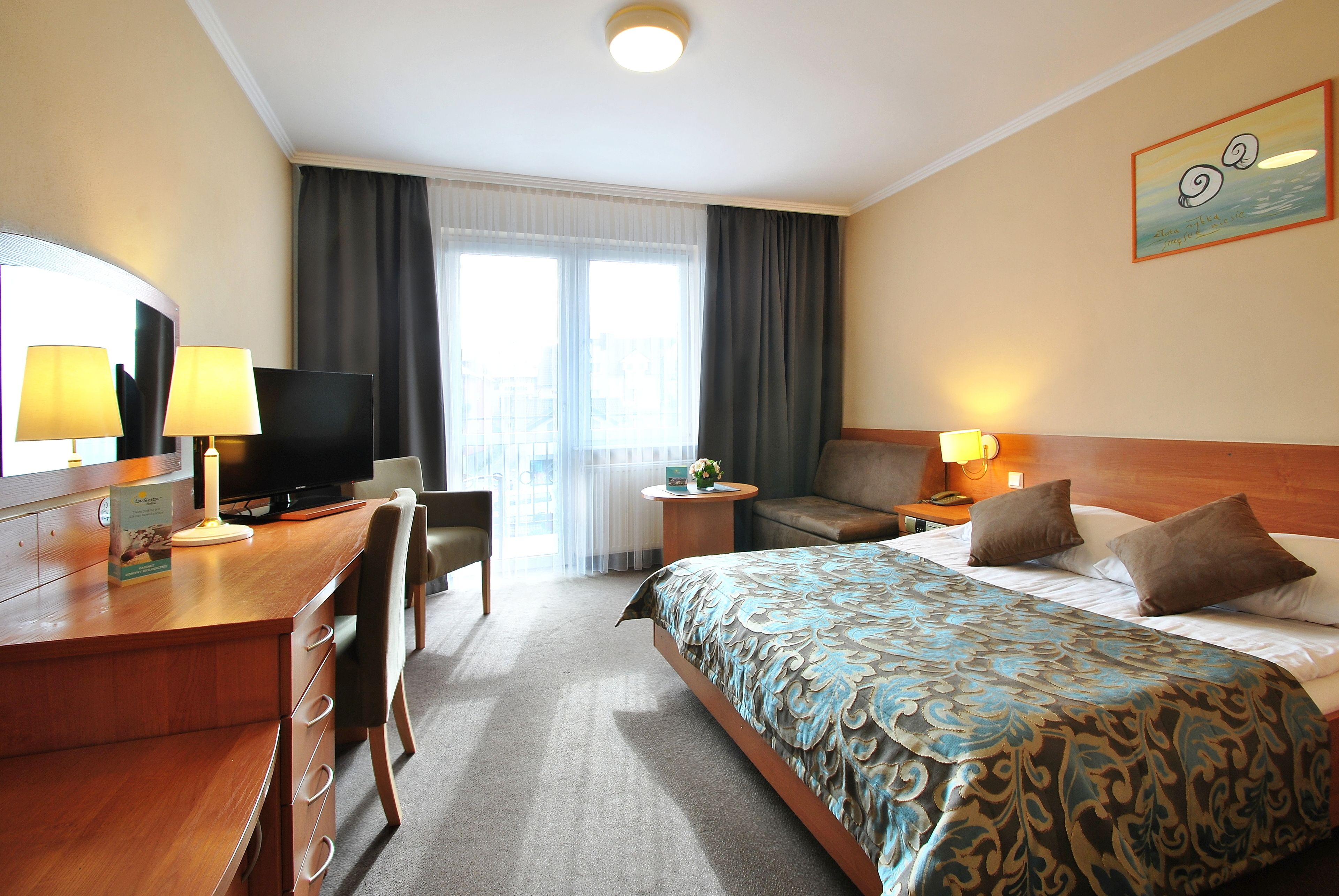 Quarto de hotel com cama, escrivaninha, cadeira, televisão e vista da cidade pela janela.