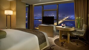 A hotel room with a large bed, a desk with a TV, a view of a cityscape at night, and a window with curtains. - Expo Center Hotel  (Jinhua)