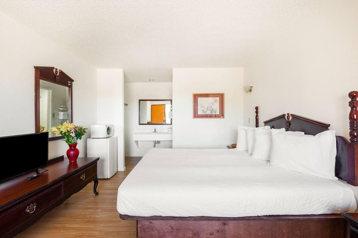 Ein Hotelzimmer mit einem großen Bett, einem Fernseher auf einer Holzkommode, einer weißen Kommode, einem Spiegel und einem durch eine offene Tür einsehbaren Badezimmer.