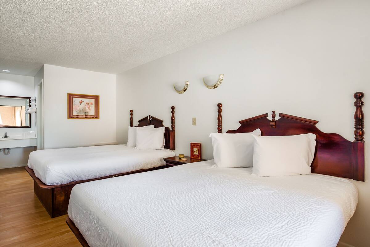 Ein Hotelzimmer mit zwei Betten, Holz-Kopfteilen, weißer Bettwäsche, einem gerahmten Bild an der Wand und einem kleinen Nachttisch mit Lampe.