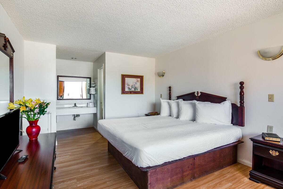 Ein Hotelzimmer mit einem hölzernen Bett, einer roten Vase mit Blumen, einem Fernseher und einem im Hintergrund sichtbaren Badezimmer.
