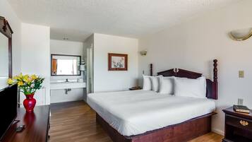 Una habitación de hotel con una cama de madera, un jarrón rojo con flores, un televisor y un baño a simple vista.