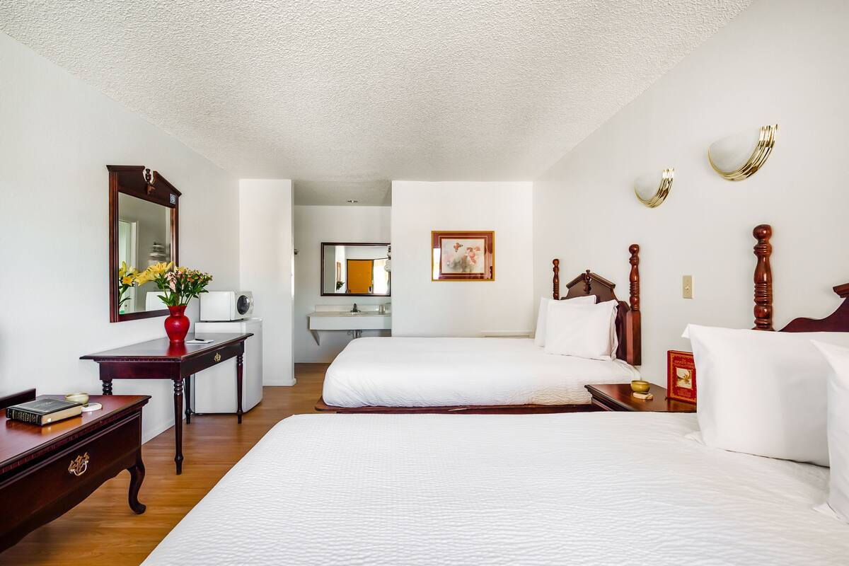 Ein Hotelzimmer mit zwei Betten, einem Schreibtisch, einem Spiegel und einem kleinen Tisch mit einer Vase.